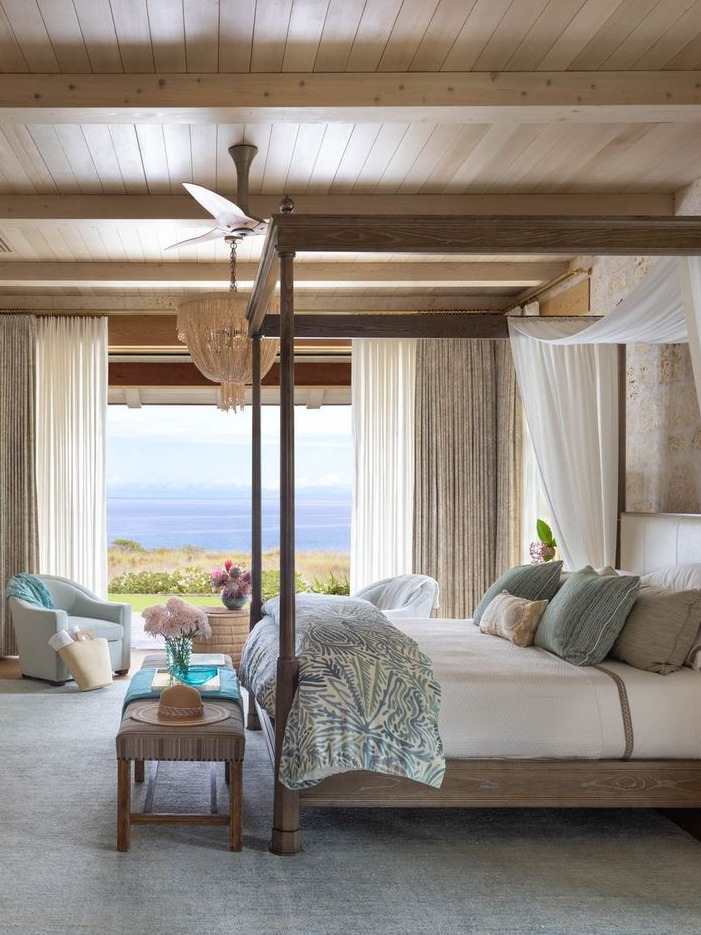 A luxurious bedroom featuring an elegant four-poster bed adorned with crisp white linens and decorative pillows in soft shades of blue and green. The room is beautifully illuminated by natural light streaming through large windows, offering a stunning view of the landscape. Sheer curtains add a touch of softness, while a stylish chandelier provides a focal point above. A cozy seating area with comfortable chairs and a small table enhances the inviting atmosphere. This sophisticated space combines classic design elements with modern comforts, creating a perfect retreat for relaxation.