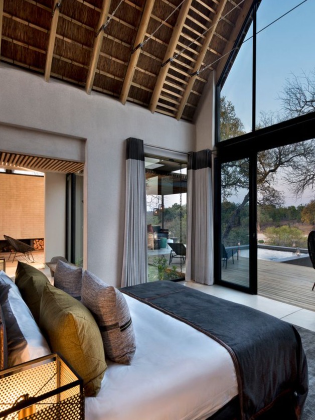 A stunning bedroom featuring a modern design with a plush bed dressed in white linens and rich green and gray decorative pillows. The room boasts a dramatic thatched ceiling and expansive glass walls that offer breathtaking views of the outdoors. Large sliding doors lead to a private deck, enhancing the connection to nature. Stylish bedside lamps and a unique side table add elegant touches, creating a serene and inviting atmosphere perfect for relaxation.