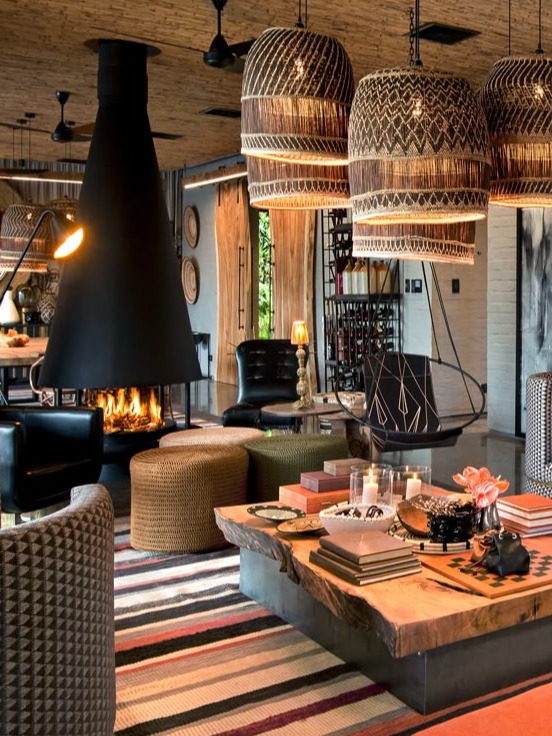 A vibrant and contemporary living room featuring a striking black fireplace as the focal point, surrounded by eclectic seating options. The space is adorned with large woven pendant lights that add texture and warmth. A unique coffee table made of natural wood displays colorful books and decorative items, while a bold striped rug ties the room together. The combination of modern and traditional elements creates an inviting atmosphere, perfect for socializing and relaxation.