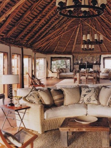 A charming and rustic living area in a safari lodge featuring a cozy sofa adorned with patterned cushions. The room boasts a stunning thatched ceiling, enhancing the natural aesthetic. Large windows allow ample light and provide views of the surrounding landscape. A wooden coffee table sits in front, accompanied by a stylish side table, creating an inviting space for relaxation and social gatherings. The warm tones and textures create a welcoming atmosphere that reflects the essence of lodge living.
