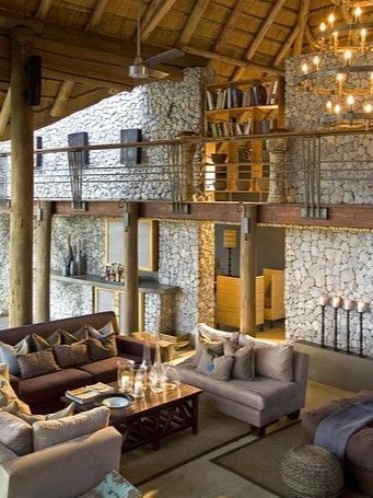 An elegant living space in a luxury lodge featuring a striking stone wall that adds texture and character to the room. Plush sofas in soft hues are arranged around a wooden coffee table, inviting relaxation. A modern chandelier hangs from the high thatched ceiling, enhancing the open feel of the space. A stylish bookshelf is visible in the background, further contributing to the sophisticated ambiance. The overall design blends natural materials with contemporary elements, creating a warm and inviting atmosphere.