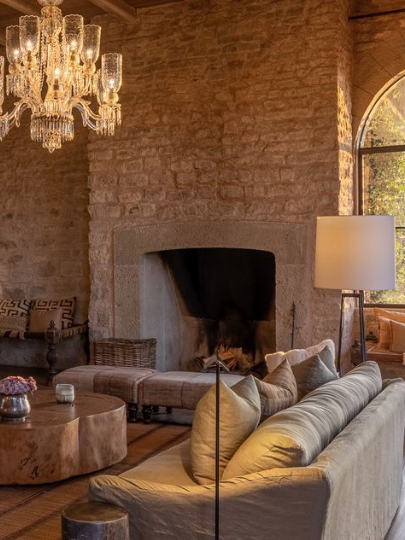 An elegant living room featuring a combination of rustic stone walls and modern decor. A stunning chandelier hangs from the wooden ceiling, illuminating the cozy space, which includes plush seating and a warm fireplace. Large arched windows provide natural light and beautiful views, creating a serene and inviting atmosphere perfect for relaxation.