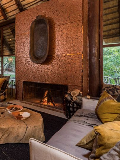 A cozy safari lodge living room featuring a striking stone fireplace and a high, thatched ceiling. The space is furnished with comfortable sofas adorned with colorful cushions and a unique wooden coffee table. Large windows frame views of the surrounding nature, creating a warm and inviting atmosphere perfect for relaxation and gathering by the fire.