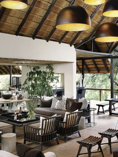 A chic safari lodge living area featuring a spacious layout with high thatched ceilings and modern pendant lighting. The room is elegantly furnished with black and white decor, including stylish seating and decorative elements. Large windows provide stunning views of the surrounding nature, creating a bright and inviting atmosphere for relaxation and socializing.